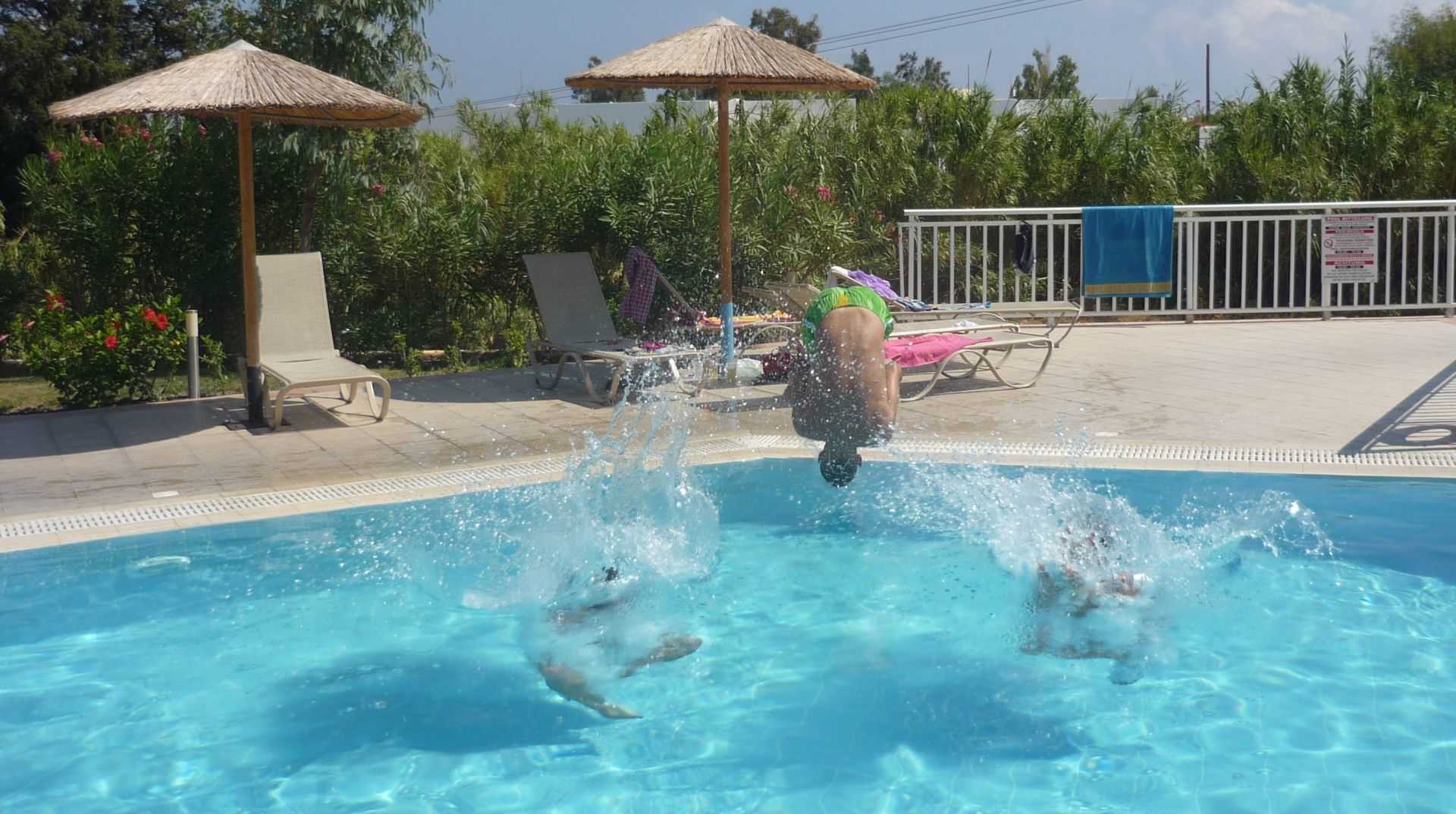 Ludzie skaczący do basenu, w tle leżaki i parasolebasenowe.